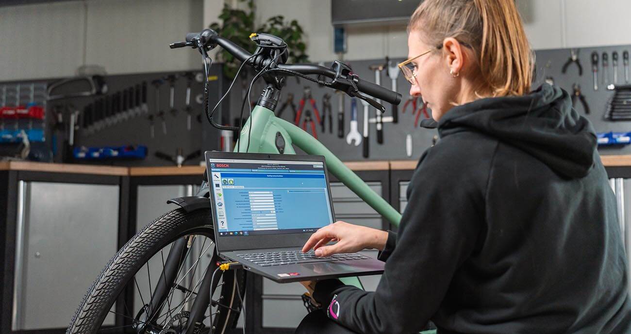 Blonde Frau mit Brille arbeitet in Fahrradwerkstatt am Laptop, um ein Update des E-Bike-Motors vorzunehmen