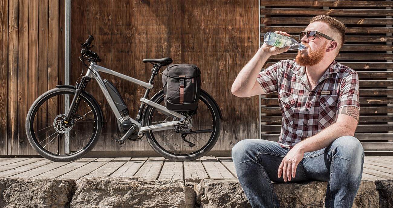 Mann im karierten Hemd sitzt auf einer Stufe und trinkt aus einer Wasserflasche. Hinter ihm steht ein Fahrrad mit Gepäckträgertasche