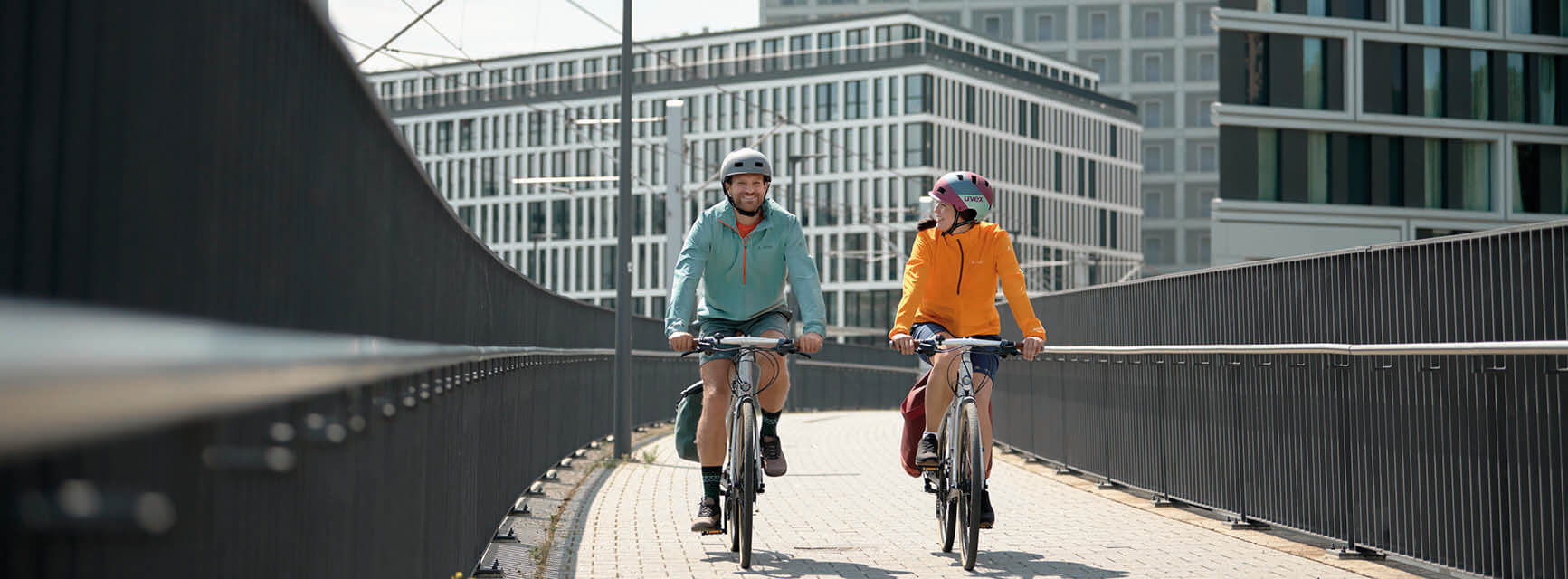 Ein Mann und frau sind bei Sonnenschein mit dem Fahrrad unterwegs. Beide lachen und tragen einen Helm.