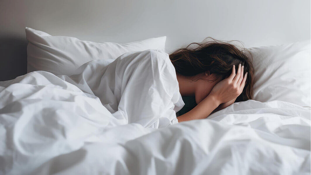 Eine Frau mit braunen Haaren liegt im Bett und vergräbt ihr Gesicht unter ihren Händen.