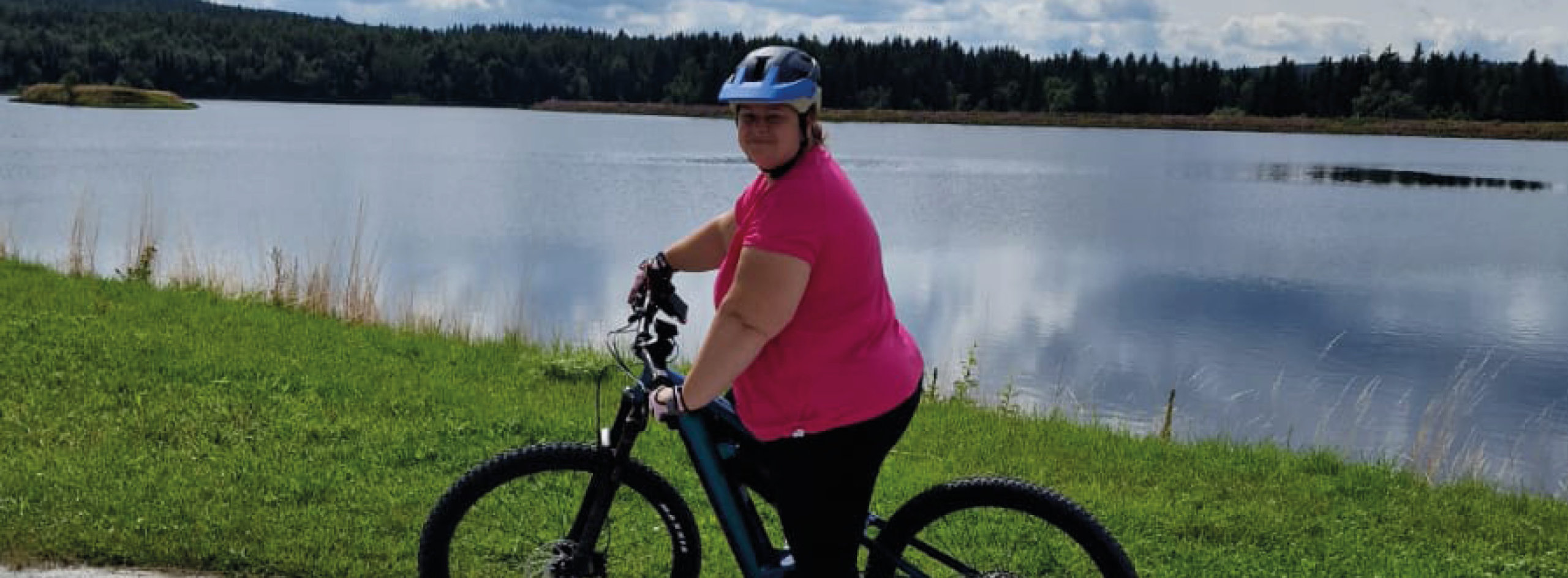 Eine Frau mit pinkem Shirt und blauem Helm steht mit ihrem E-Bike vor einem See und lächelt in die Kamera