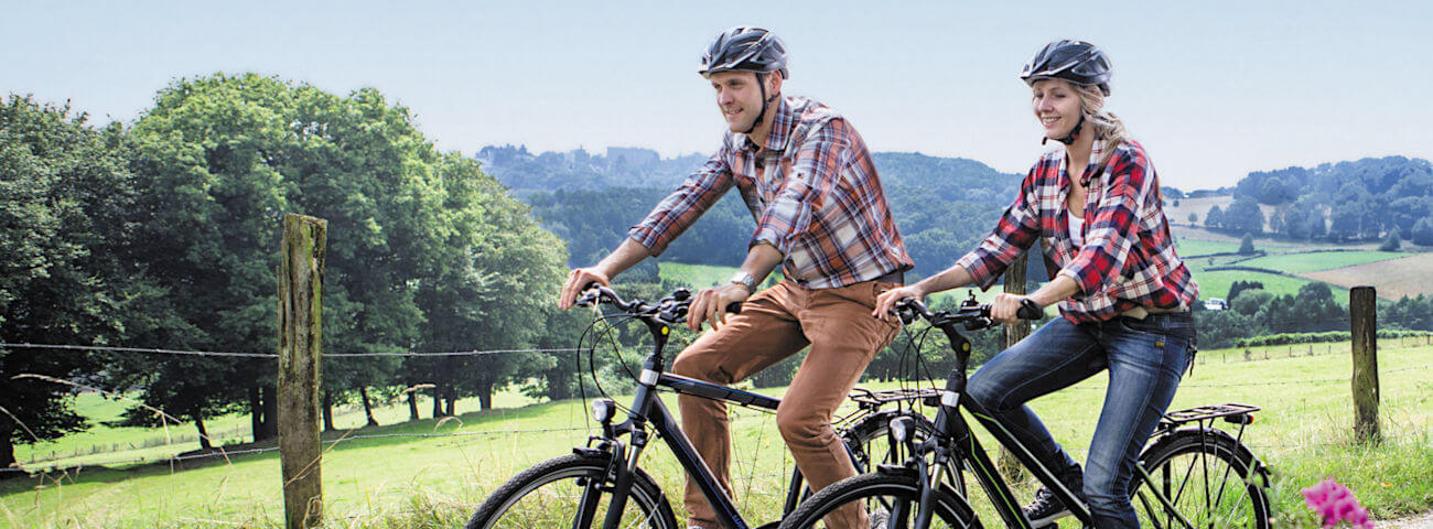 Zwei Radfahrer mit Helm fahren auf einem Weg durch eine grüne Landschaft