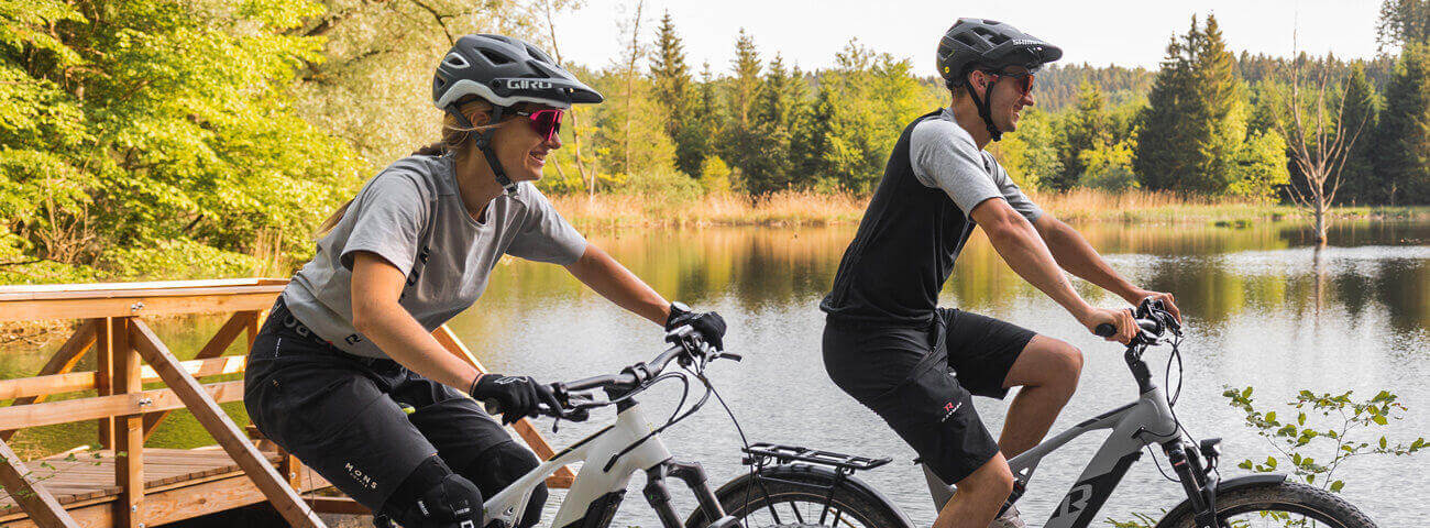 Zwei Radfahrer fahren mit E-Mountainbikes von R Raymon an einem See vorbei