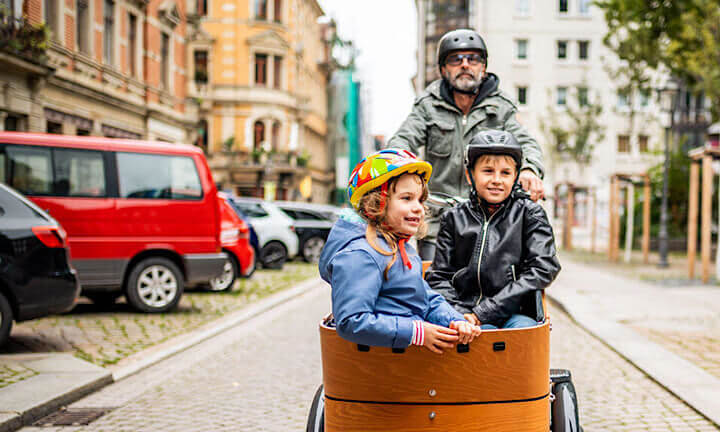 Papa in Rocker-Outfit fährt zwei Kinder auf Lastenrad durch die Stadt
